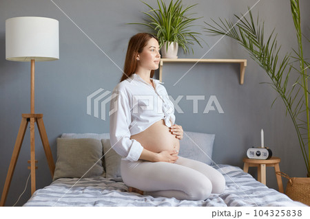 Beautiful gentle brown haired pregnant woman with bare belly wearing white clothing sitting in bed in light bedroom, touching her tummy, looking away, thinking about expecting baby. 104325838