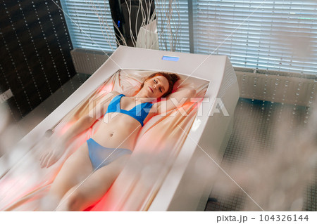 High-angle view of relaxed woman in swimsuit lying with closed eyes on electric massage bed, having rest enjoying spa procedure at luxury spa salon. Sensual redhead female enjoying wellness weekend. 104326144