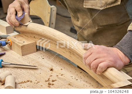 Carpenter sands bending wooden railing with sandpaper in workshop closeup. Senior master makes detail of spiral staircase for home interior Carpenter sands bending wooden railing with sandpaper in workshop closeup. Senior master makes detail of spiral staircase for home interior 104326248