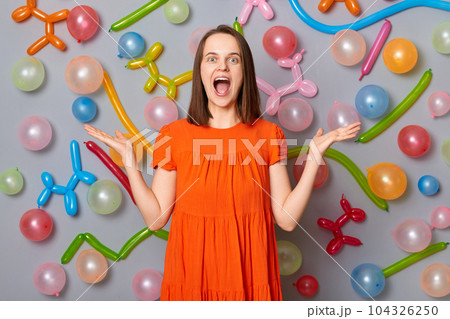 Overjoyed female. Surprised emotion. Excited woman with brown hair wearing orange dress raised her arms screaming wow standing against gray wall decorated with colorful balloons. Overjoyed female. Surprised emotion. Excited woman with brown hair wearing orange dress raised her arms screaming wow standing against gray wall decorated with colorful balloons. 104326250