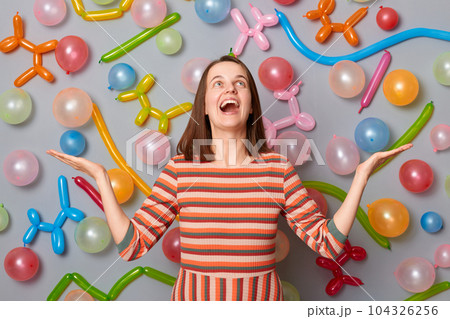 Extremely happy overjoyed woman wearing striped dress standing against gray wall with balloons decoration looking up celebrating screaming with happiness and joy. Extremely happy overjoyed woman wearing striped dress standing against gray wall with balloons decoration looking up celebrating screaming with happiness and joy. 104326256