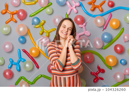 Cute woman with brown hair wearing striped dress looking at camera with charming smile expressing happiness standing against gray wall decorated with colorful balloons. 104326260
