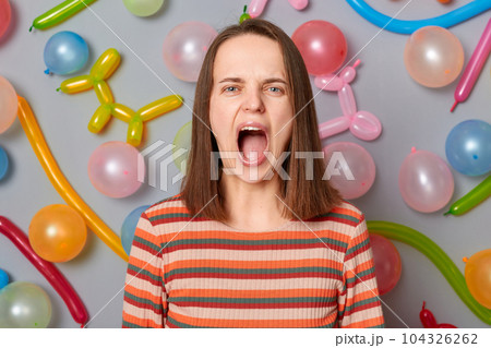 Angry despair woman wearing striped dress standing against gray wall with balloons decoration screaming with hate and aggression looking at camera open mouth. Angry despair woman wearing striped dress standing against gray wall with balloons decoration screaming with hate and aggression looking at camera open mouth. 104326262
