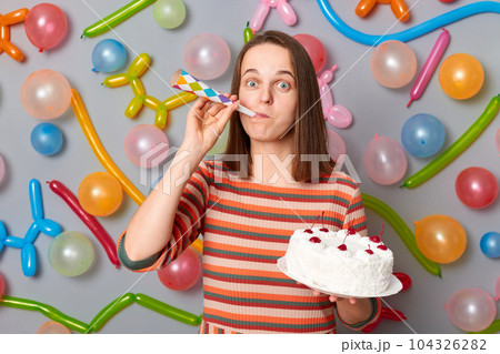 Festive funny happy woman with brown hair wearing striped dress holding cake blowing party horn having fun on party standing against gray wall decorated with colorful balloons. 104326282