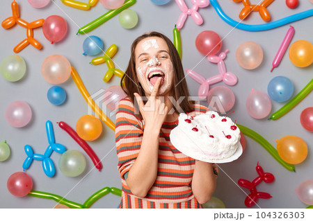 happy smiling woman with cake cream on face wearing striped dress standing against gray wall decorated with colorful balloons licking finger with birthday dessert. happy smiling woman with cake cream on face wearing striped dress standing against gray wall decorated with colorful balloons licking finger with birthday dessert. 104326303