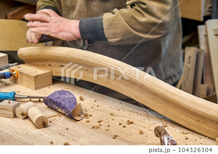 Carpenter sands bending wooden railing with sandpaper in workshop closeup. Senior master makes detail of spiral staircase for home interior Carpenter sands bending wooden railing with sandpaper in workshop closeup. Senior master makes detail of spiral staircase for home interior 104326304