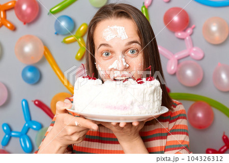Positive happy beautiful woman in striped dress standing against gray wall decorated with colorful balloons biting her birthday cake after dipping face in cream. 104326312