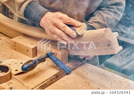 Carpenter sands bending wooden railing with sandpaper in workshop closeup. Senior master makes detail of spiral staircase for home interior 104326341