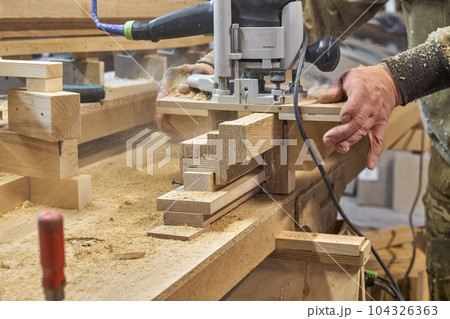 Carpenter milling wooden handrail with router manual milling machine in workshop close up. Handyman processes timber piece with electric instrument Carpenter milling wooden handrail with router manual milling machine in workshop close up. Handyman processes timber piece with electric instrument 104326363