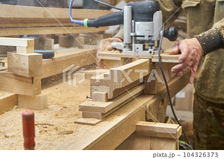 Carpenter milling wooden handrail with router manual milling machine in workshop close up. Handyman processes timber piece with electric instrument Carpenter milling wooden handrail with router manual milling machine in workshop close up. Handyman processes timber piece with electric instrument 104326373
