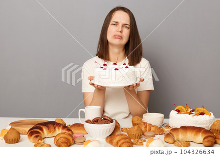 Sad upset attractive woman wearing white T-shirt isolated over gray background sitting at festive table among homemade desserts holding showing tasty cake. 104326634