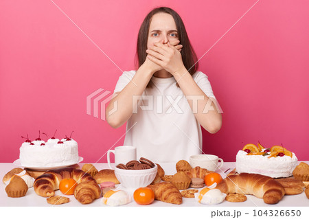 Overeating sad brown haired woman in white t shirt sitting at table isolated over pink background feeling nausea after eating sugary cakes. Overeating sad brown haired woman in white t shirt sitting at table isolated over pink background feeling nausea after eating sugary cakes. 104326650