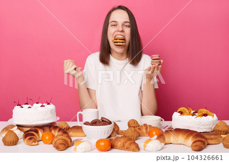 Extremely hungry brown haired woman in white t shirt breaking diet sitting at table with desserts eating different sweets isolated over pink background. 104326661