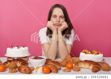 Bored girl posing at table with sugary desserts and bakery isolated over pink background looking at camera with unhappy face keeps hands under chin. Bored girl posing at table with sugary desserts and bakery isolated over pink background looking at camera with unhappy face keeps hands under chin. 104326681