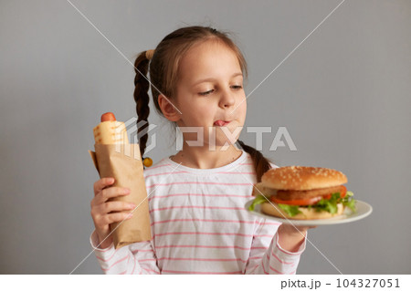 Hungry funny cute little girl with pigtails with hot dogs and hamburger isolated over gray background choosing snack for dinner showing tongue out. 104327051