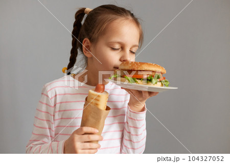 Charming cute beautiful little girl with pigtails with hot dog smelling aromat fresh baked hamburger standing with closed eyes isolated over gray background 104327052