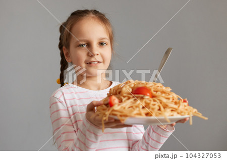 Cute smiling little kid girl with pigtails eating spaghetti holding big plate with pasta looking at camera waiting for dinner being extremely hungry. 104327053