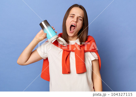 Tired exhausted woman wearing white t-shirt and jumper over neck standing isolated over blue background being sleepless showing empty thermos with no coffee. Tired exhausted woman wearing white t-shirt and jumper over neck standing isolated over blue background being sleepless showing empty thermos with no coffee. 104327115