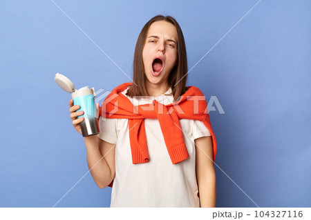 Sleepy yawning woman wearing white t-shirt and jumper over neck standing isolated over blue background holding thermos with coffee needs energy. Sleepy yawning woman wearing white t-shirt and jumper over neck standing isolated over blue background holding thermos with coffee needs energy. 104327116