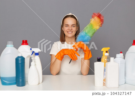 Joyful happy smiling woman posing at workplace with cleaning detergents sitting with pp duster in hands cleaning her house isolated over gray background. Joyful happy smiling woman posing at workplace with cleaning detergents sitting with pp duster in hands cleaning her house isolated over gray background. 104327147
