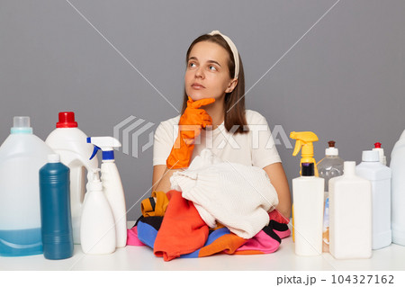Pensive thoughtful woman posing at workplace with cleaning detergents isolated over gray background holding chin looking away thinking about household tasks. 104327162