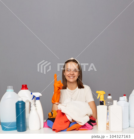 Delighted satisfied woman posing at workplace with cleaning detergents isolated over gray backgroun doing laundry pointing up at copy space for promotion. 104327163