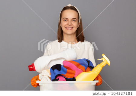 Routine washing keeps garments fresh. Housekeeper maintains clean and tidy house. Smiling cheerful woman wearing whote t-shirt with laundry bowl and detergent isolated over gray background 104327181