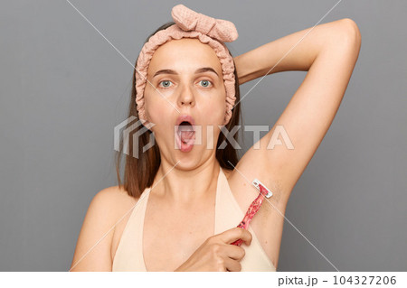Amazed surprised young woman with hair band using razor to remove hair from her armpit talking care of her beauty and hygiene posing isolated over gray background. 104327206