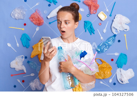 Reduce garbage pollution. Cleaning up planet. Shocked surprised woman wearing white shirt isolated on blue wall with garbage around looking at cell phone screen with open mouth. Reduce garbage pollution. Cleaning up planet. Shocked surprised woman wearing white shirt isolated on blue wall with garbage around looking at cell phone screen with open mouth. 104327506