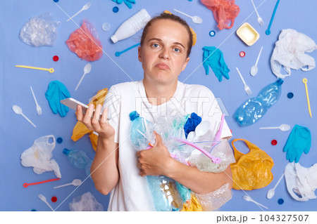 Uncertain woman wearing white shirt isolated on blue wall with garbage around standing with litter and smart phone in hands looking at camera shrugging shoulders. Uncertain woman wearing white shirt isolated on blue wall with garbage around standing with litter and smart phone in hands looking at camera shrugging shoulders. 104327507
