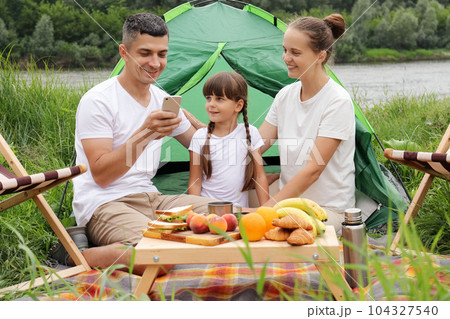 Family at riverside near the tent. Using smartphone in nature. Resting vacation outdoor. Family with little daughter enjoying time in part near the river. Family at riverside near the tent. Using smartphone in nature. Resting vacation outdoor. Family with little daughter enjoying time in part near the river. 104327540