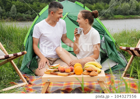 Leisure outdoor. Resting in nature. Camping near river. Happy laughing woman and man wearing casual clothing sitting at tent on ground, communicating, enjoying vacation. Leisure outdoor. Resting in nature. Camping near river. Happy laughing woman and man wearing casual clothing sitting at tent on ground, communicating, enjoying vacation. 104327556