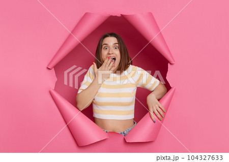 Surprised astonished brunette woman wearing striped top looking through ripped pink paper hole being impressed covering mouth with palm sees something shocked. Surprised astonished brunette woman wearing striped top looking through ripped pink paper hole being impressed covering mouth with palm sees something shocked. 104327633