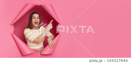 Excited positive brunette woman wearing striped top looking through ripped pink paper hole pointing at advertisement area saying wow empty space for promotion. Excited positive brunette woman wearing striped top looking through ripped pink paper hole pointing at advertisement area saying wow empty space for promotion. 104327640