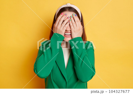 Smiling woman wearing green jacket posing isolated over yellow background hiding her eyes covering face with palms positive emotions. 104327677