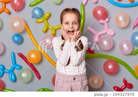 Portrait of amazed extremely happy birthday little girl with braids wearing casual clothing standing against gray wall with colorful balloons, being pleasantly surprised. Portrait of amazed extremely happy birthday little girl with braids wearing casual clothing standing against gray wall with colorful balloons, being pleasantly surprised. 104327870