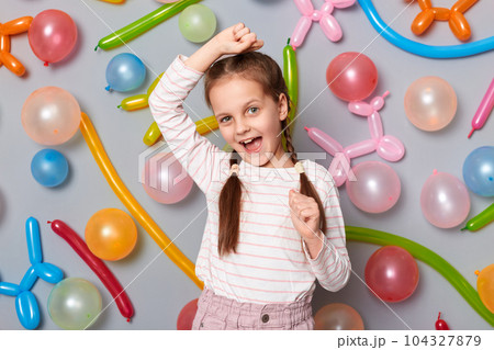 Photo of excited joyful little girl with braids wearing casual clothing clenched fists raised arms celebrating birthday dancing standing against gray wall with colorful balloons. 104327879