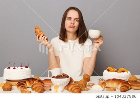 Photo of sad upset woman with brown hair sitting at table with delicious pastry, being overeating, holding cup and croissant in hands, isolated over gray background. 104327986