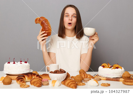 Portrait of shocked surprised woman with brown hair sitting at table isolated over gray background, looking at camera with big eyes, holding cup of tea and bagel in hands, saying wow. Portrait of shocked surprised woman with brown hair sitting at table isolated over gray background, looking at camera with big eyes, holding cup of tea and bagel in hands, saying wow. 104327987