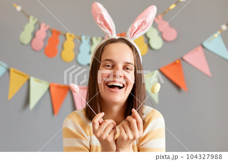 Happy Easter. Charming smiling woman with dark hair wearing rabbit ears holding delicious Easter cake pops, laughing, looking at camera, isolated on gray decorated background Happy Easter. Charming smiling woman with dark hair wearing rabbit ears holding delicious Easter cake pops, laughing, looking at camera, isolated on gray decorated background 104327988