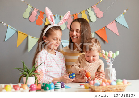Portrait of smiling mother sitting at table with her daughters and painting eggs, little girl wearing bunny ears, family being in good festive mood, posing against gray decorated wall. 104328107
