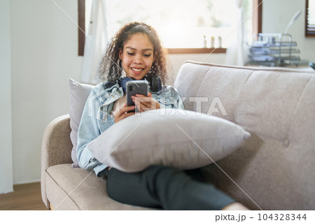 Portrait of an African American sitting on a sofa using a phone and wearing headphones to relax. 104328344