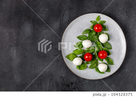 Caprese salad in the form of a Christmas tree. Festive tomato mozzarella and basil appetizer on light plate. Caprese salad in the form of a Christmas tree. Festive tomato mozzarella and basil appetizer on light plate. 104328375