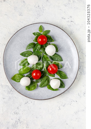Caprese salad in the form of a Christmas tree. Festive tomato mozzarella and basil appetizer on light plate. Caprese salad in the form of a Christmas tree. Festive tomato mozzarella and basil appetizer on light plate. 104328376