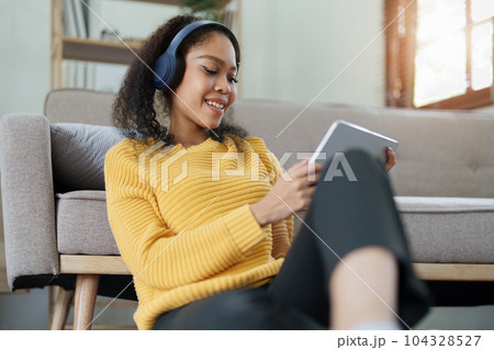 Portrait of an African American with headphones and tablet smiling happily listen music while relaxing on the sofa at home. 104328527
