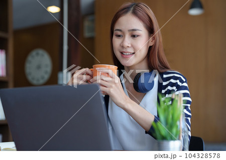 Portrait of a young, attractive Asian teenage girl using a computer and drinking coffee while studying online in the morning at the library. Portrait of a young, attractive Asian teenage girl using a computer and drinking coffee while studying online in the morning at the library. 104328578