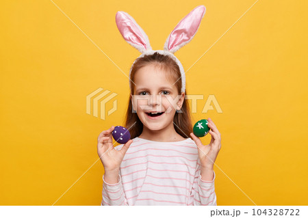 Portrait of excited charming girl with rabbit ears on her head with colored eggs in hands, being in good mood, celebrating spring holiday, posing isolated on yellow background 104328722