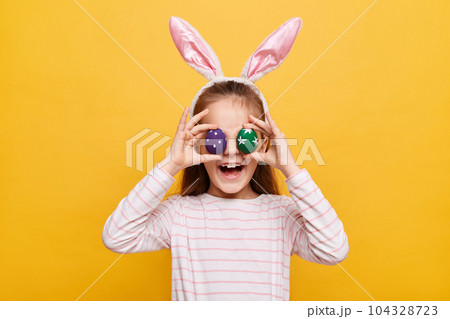 Portrait of funny little girl with rabbit ears with covering her eyes colored purple and green eggs having fun smiling happily posing isolated on yellow background 104328723