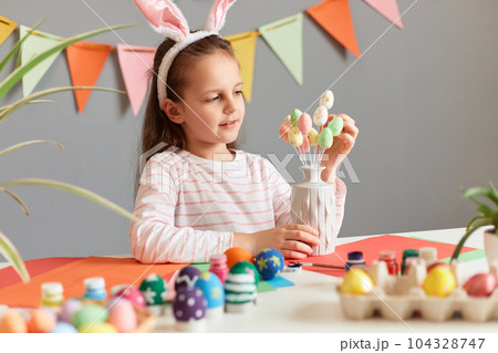 Portrait of cute dark haired little girl holding small multicolored painted dyed easter eggs on sticks, wearing bunny ears, celebrating Easter, sitting at table among colored Easter eggs. 104328747