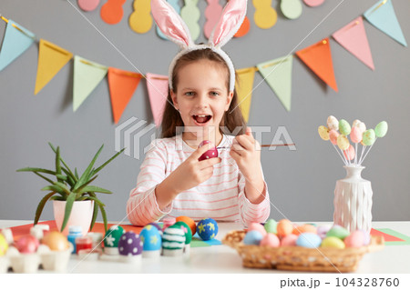 Photo of excited adorable little girl painting aster eggs with brush, wearing bunny ears looking at camera with happiness, celebrating Easter, posing against decorated gray background. 104328760